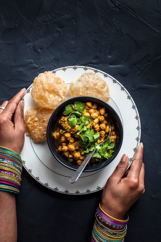 Channa nu shaak, chickpea masala in a black bowl surround by poori, puri on a plate with lady wearing colourful bangles holding it