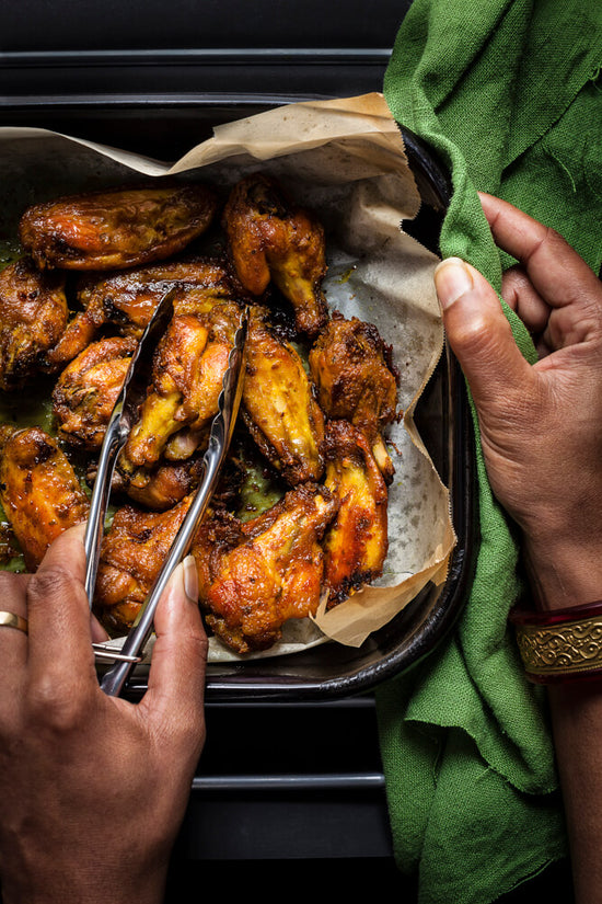 Masala chicken nibbles inside a baking tray and hand holding tongs holding one nibble
