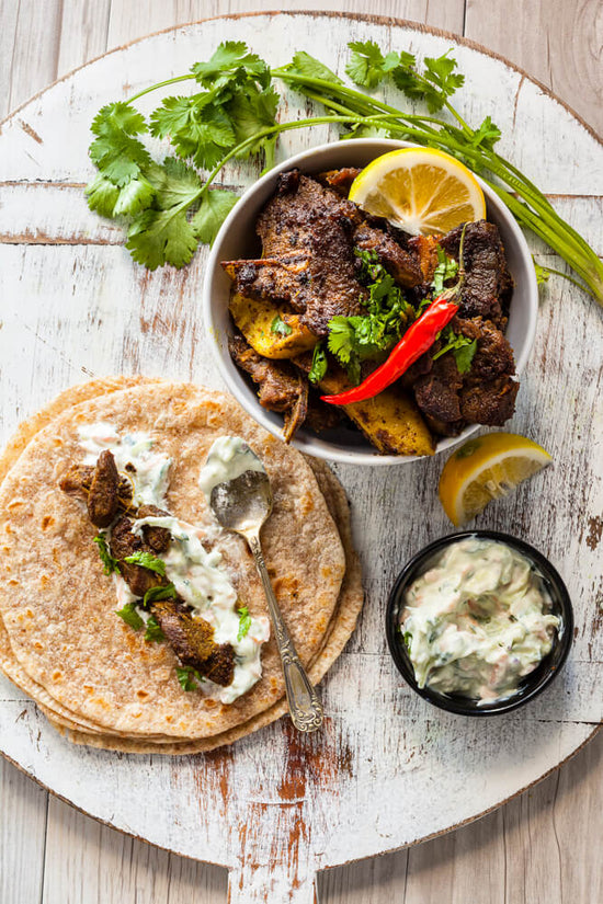 Masala Lamb with rotli and raitu on a tray with coriander and lemon slices to make masala wraps