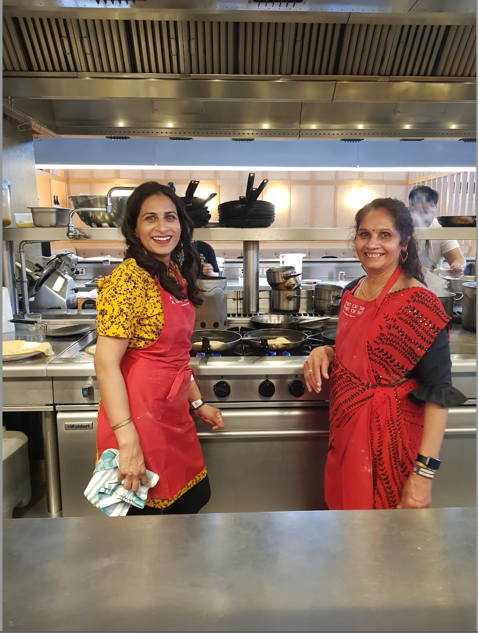 Laxmi in sari and Jayshri in dress and apron standing in commercial kitchen; share what they love about their cookbook and why it's the only thing you'll need to produce dishes for your family in an Authentic Gujarati home cooked way - the way of mum
