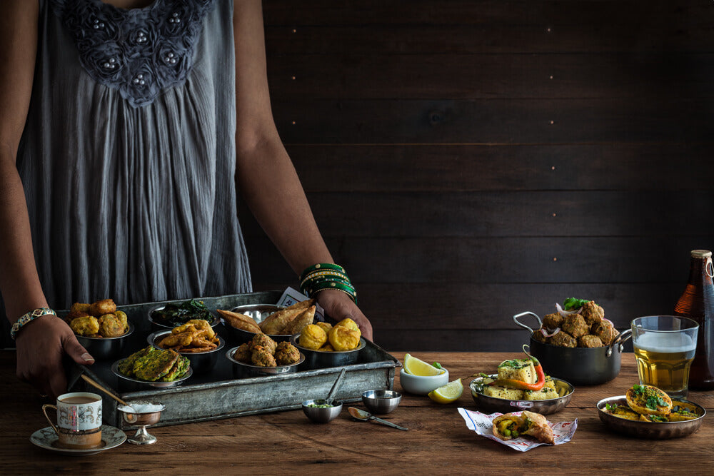 Gujarati Nasto, pakora, dhokla, samosa, bateta vada, bataka vada, curried eggs, a lady holding a selection with chai in a tea cup and beer in a mug